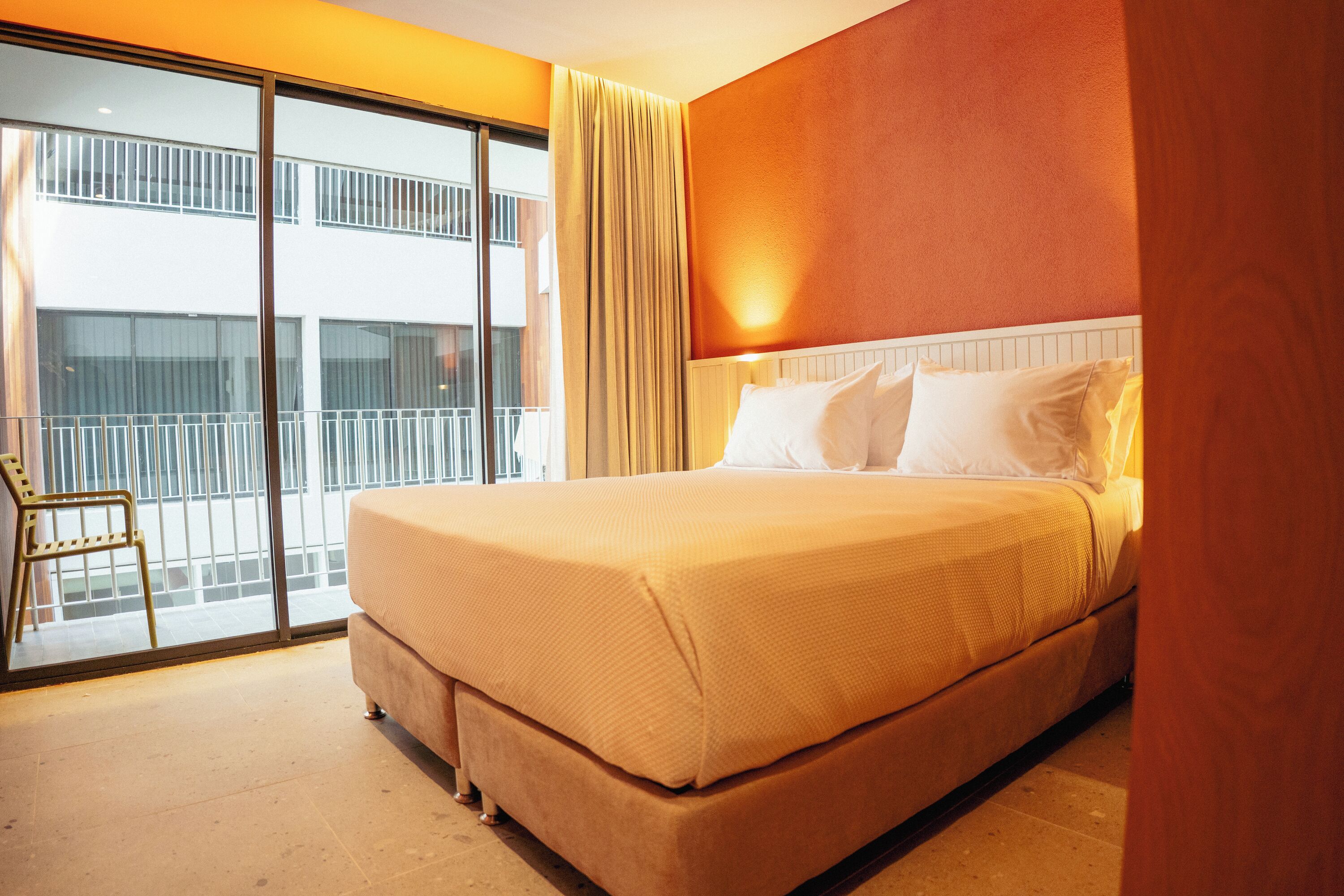 A hotel room with a large bed, a chair, a balcony with a view of buildings, and a window with curtains.