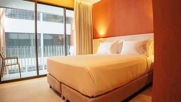 A hotel room with a large bed, a chair, a balcony with a view of buildings, and a window with curtains.