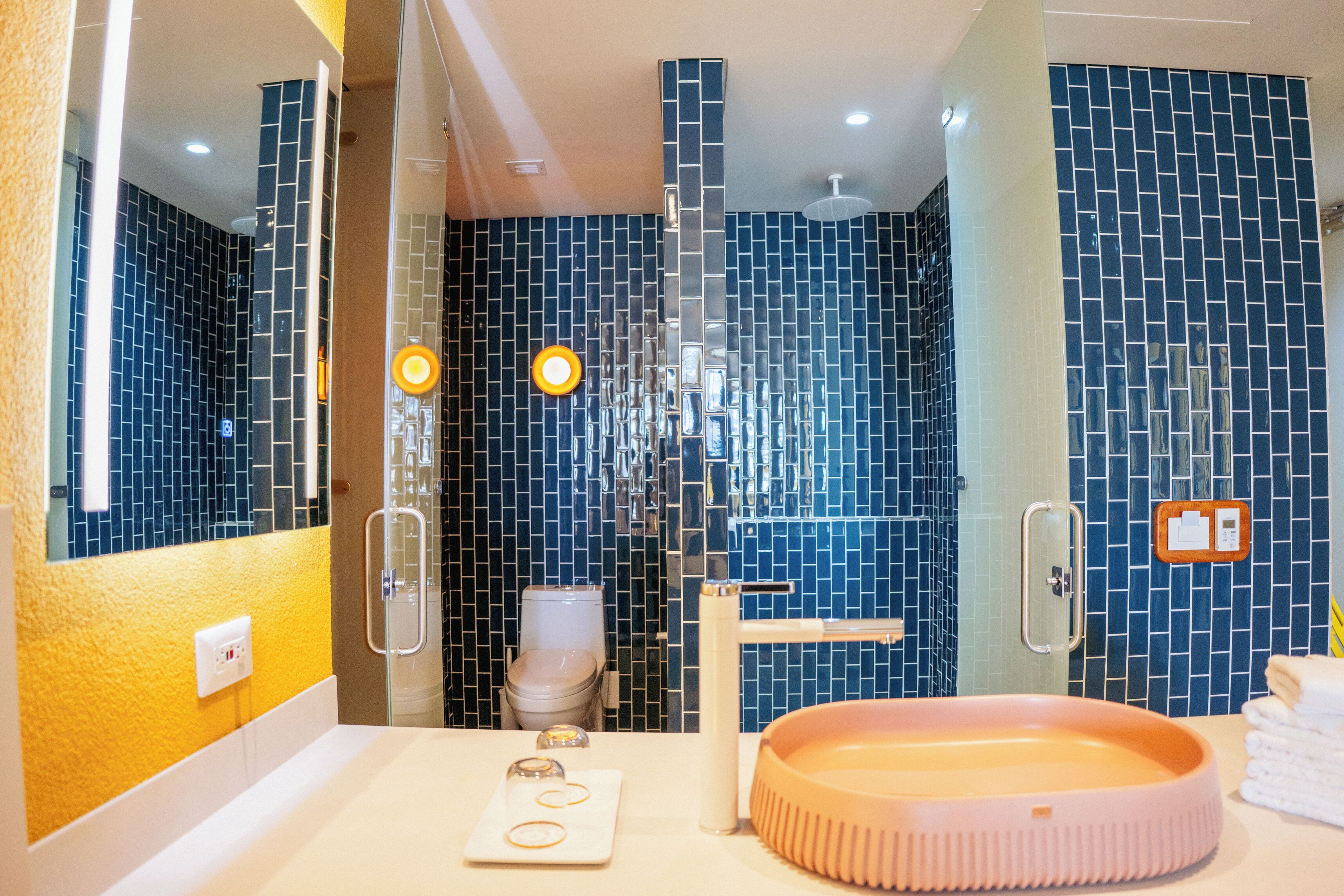 Un baño con ducha de azulejos azules, un lavabo blanco con lavamanos rosa y un espejo que refleja la habitación.