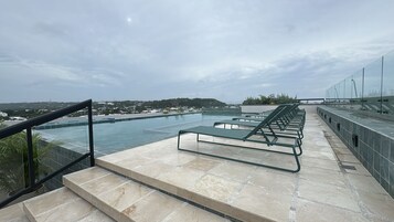 Una piscina infinita con sillones reclinables, terraza de piedra y barandilla de vidrio con vistas a un paisaje urbano.