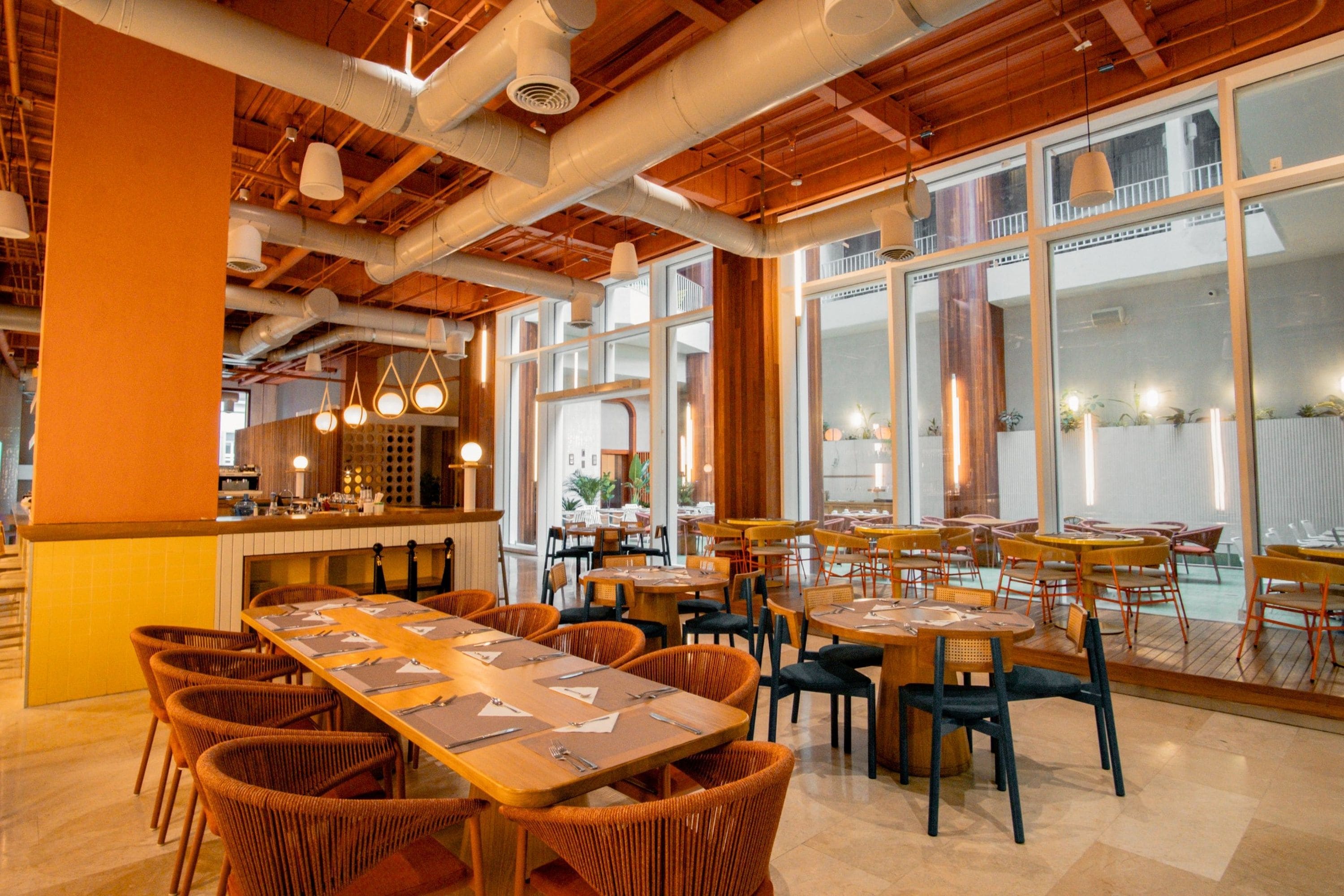 Interior de restaurante con techos altos, conductos expuestos y una combinación de asientos de madera y tapicería.