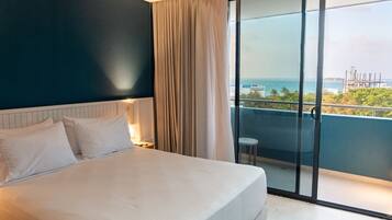 Quarto de hotel com cama, mesa de cabeceira, abajur, janela com cortinas e vista para o oceano e edifícios.