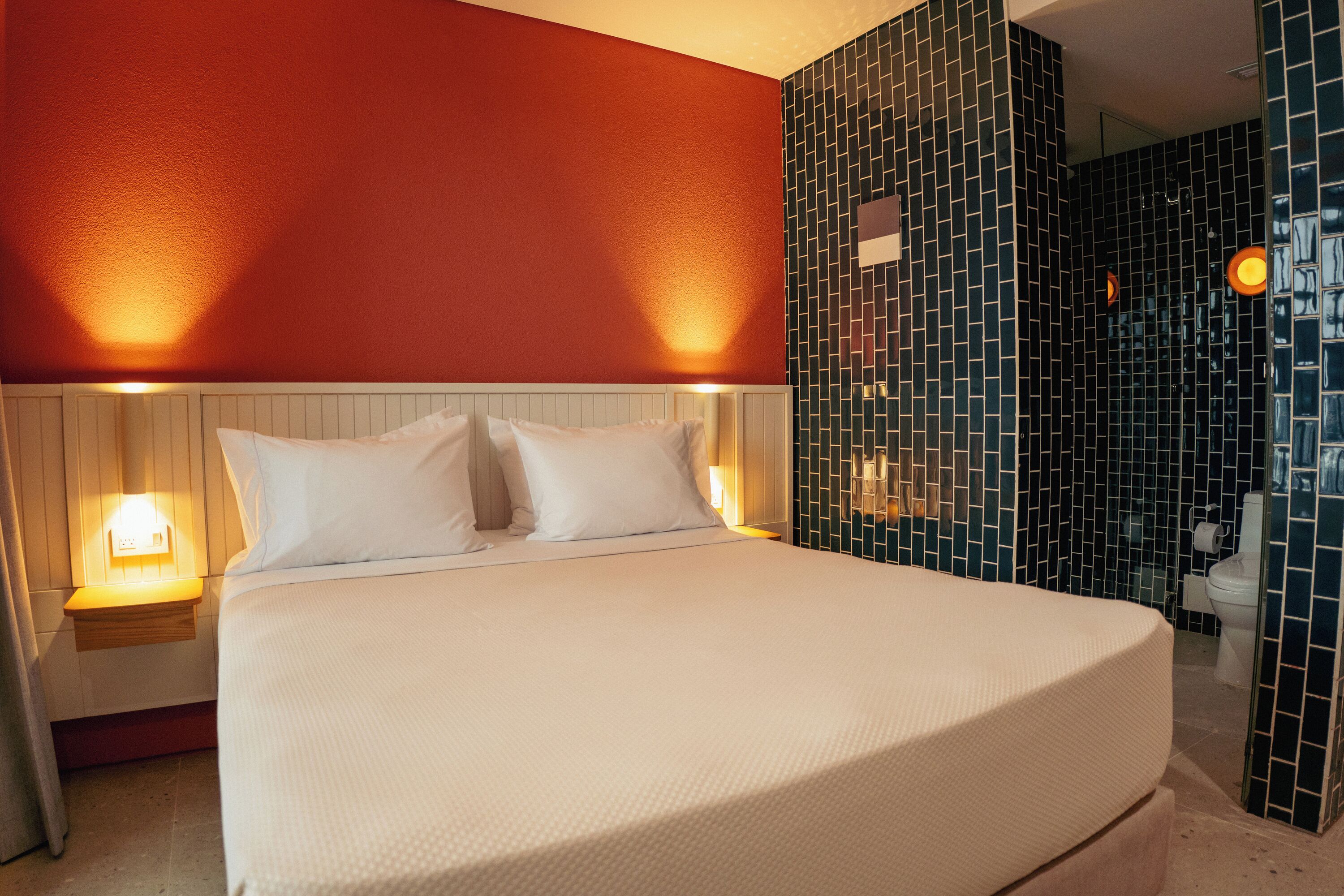 Ein Hotelzimmer mit einem großen Bett, einem Nachttisch mit Lampe und einem Badezimmer mit gefliesten Wänden.