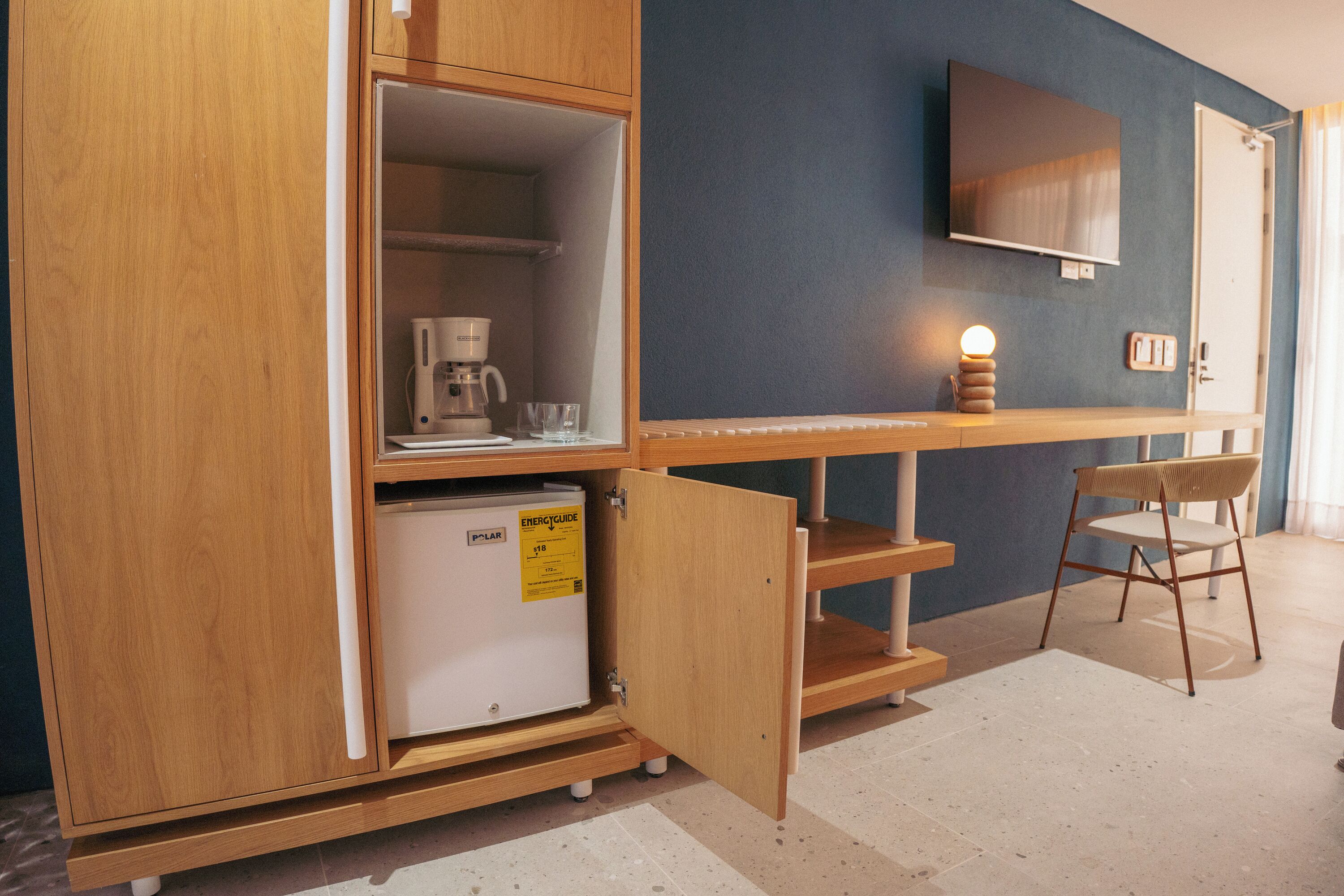 Una kitchenette compacta con cafetera, refrigerador pequeño y una silla.