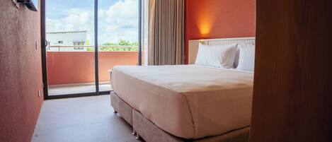 Habitación de hotel con una cama grande, puerta corrediza de vidrio a un balcón y vista al cielo y edificios.