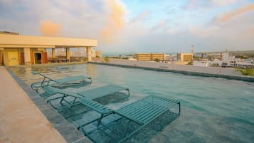 Uma piscina infinita no terraço com espreguiçadeiras, vista para a paisagem urbana e céu claro.