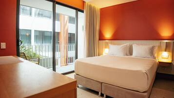 A hotel room with a bed, bedside lamps, a desk, and a view of a building through a glass door.