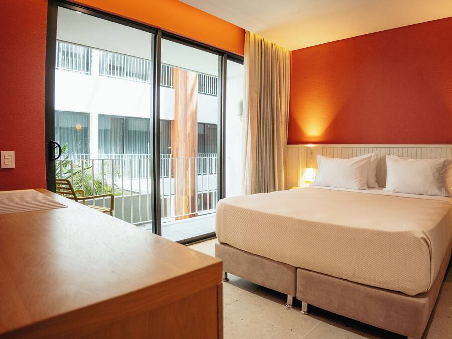 A hotel room with a bed, bedside lamps, a desk, and a view of a building through a glass door.