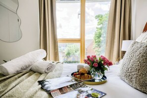 A hotel room with a bed, a window with curtains, a tray with croissants and a vase of flowers, and a magazine on the bed.