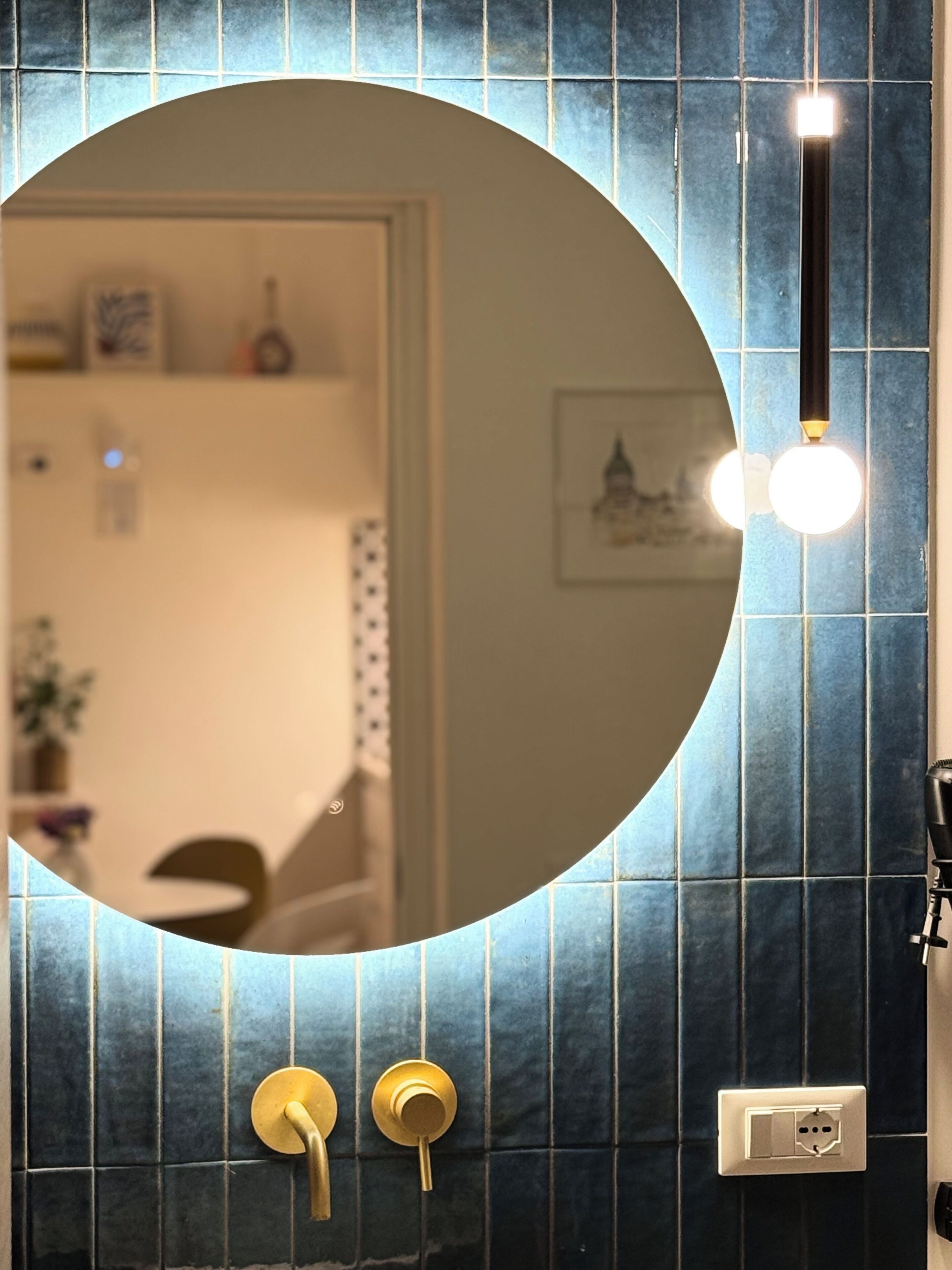 Une salle de bain avec un miroir rond, un porte-serviettes, un luminaire et une prise électrique fixée au mur.