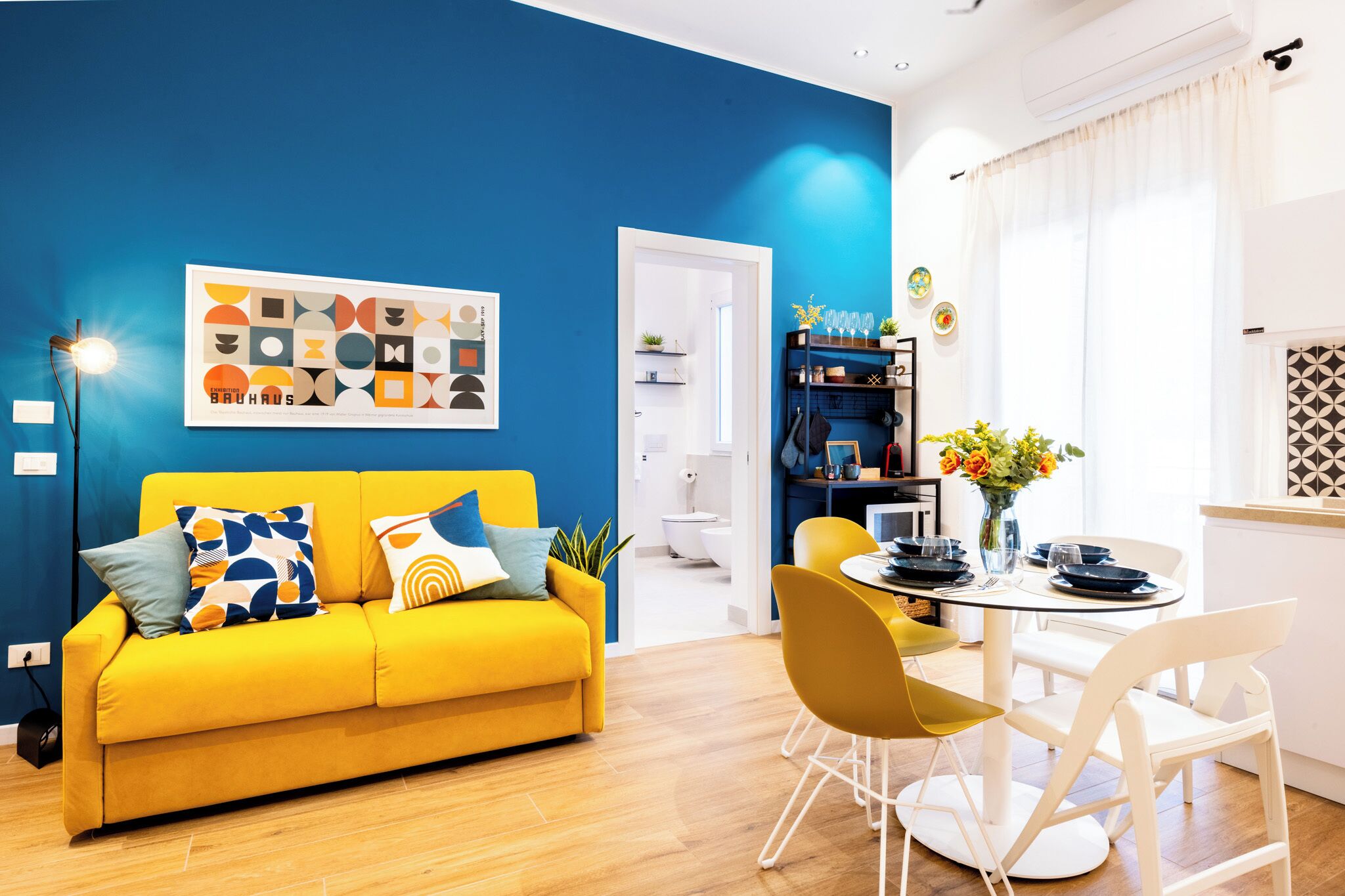 A modern living room with a yellow sofa, a dining table with chairs, and a blue accent wall.
