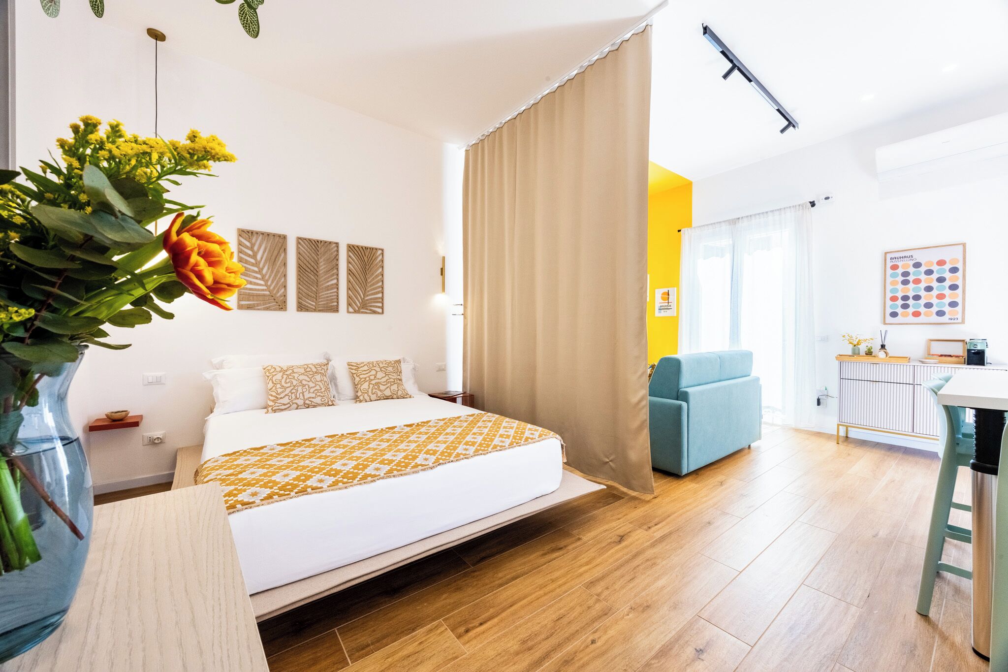A modern hotel room with a large bed, a small table, a chair, and a desk. There is a curtain, a vase with flowers, and a wall with decorative items.