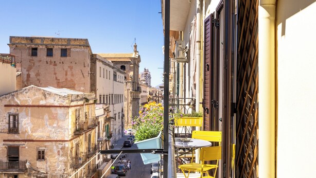 A narrow street with buildings on either side, a balcony with yellow chairs, and a potted plant with flowers. - Appartamenti Pellegrino (Palermo)