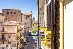 Una calle estrecha flanqueada por edificios, un balcón con sillas amarillas y una maceta con flores.