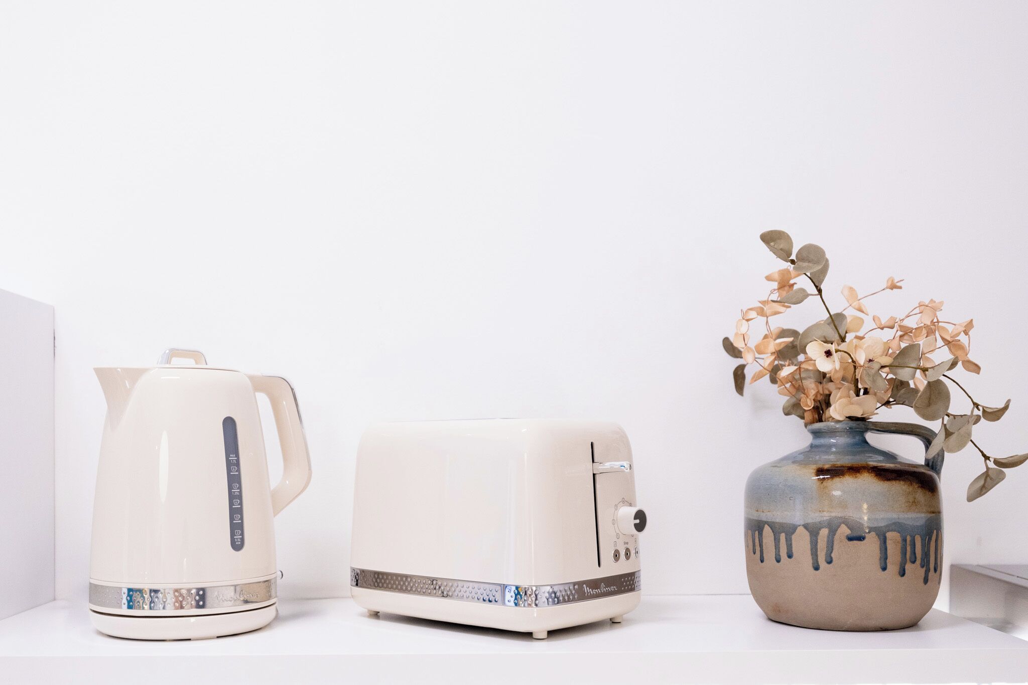 A white kettle, a white toaster, and a vase with dried flowers on a white shelf.