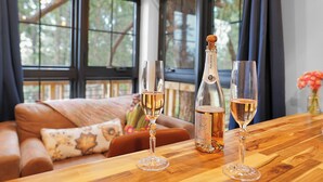 A wooden table with two glasses of champagne and a bottle of wine in a room with large windows and a cozy seating area.