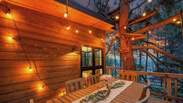 A wooden deck with a table set for dining, surrounded by chairs, and string lights.
