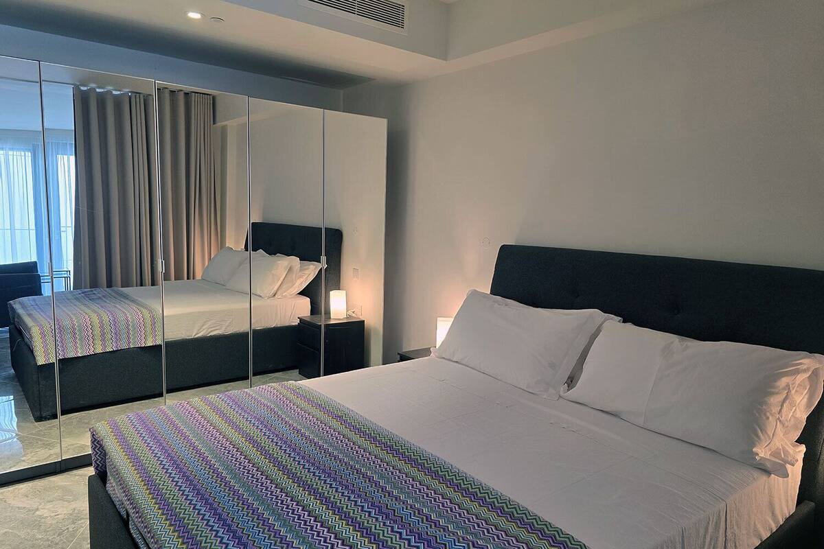 Une chambre d’hôtel moderne avec un grand lit, une table de chevet avec une lampe, un miroir et une vue sur l’extérieur par la fenêtre.