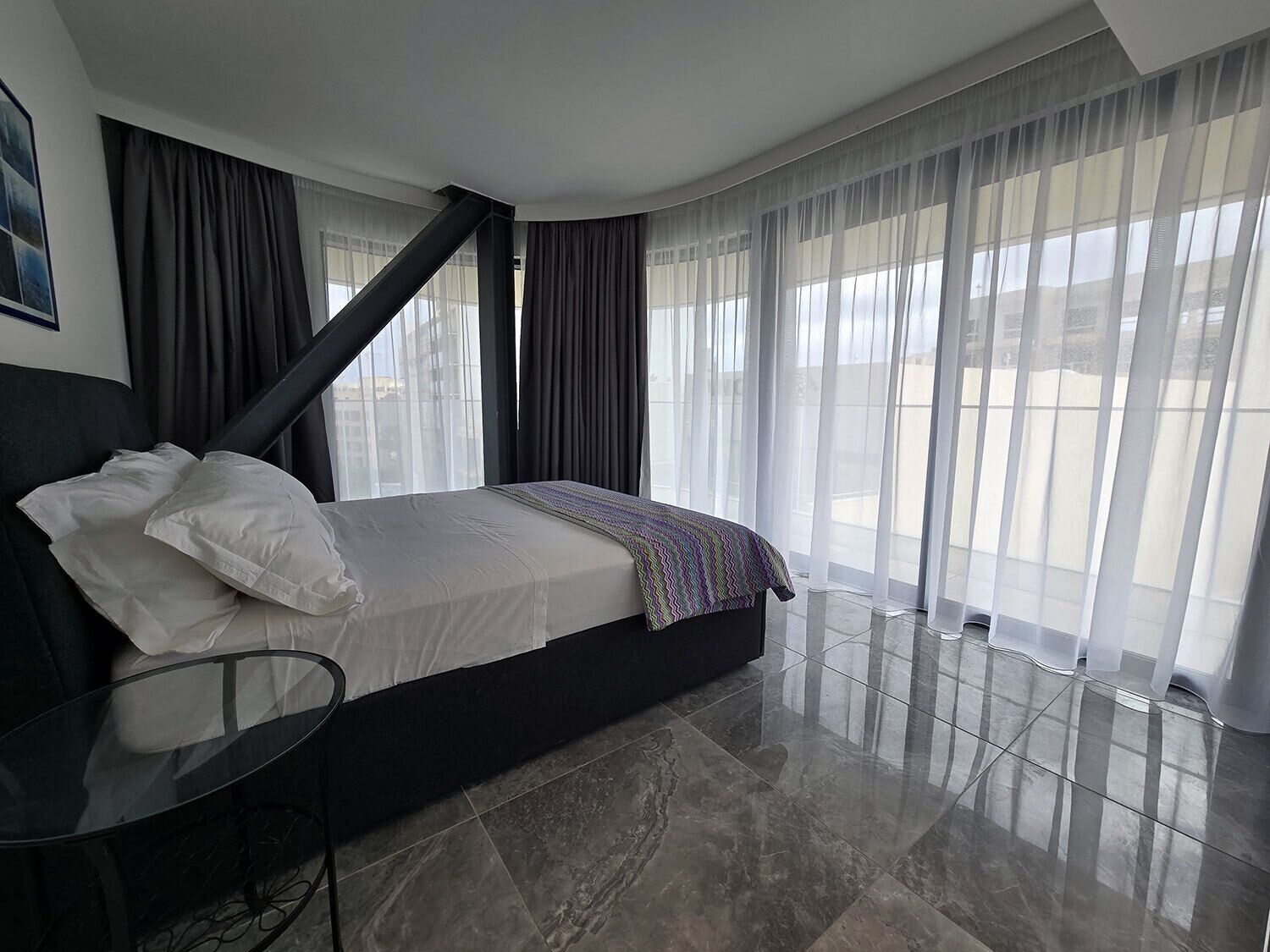 A modern hotel room with a large bed, a glass table, and large windows with sheer curtains.