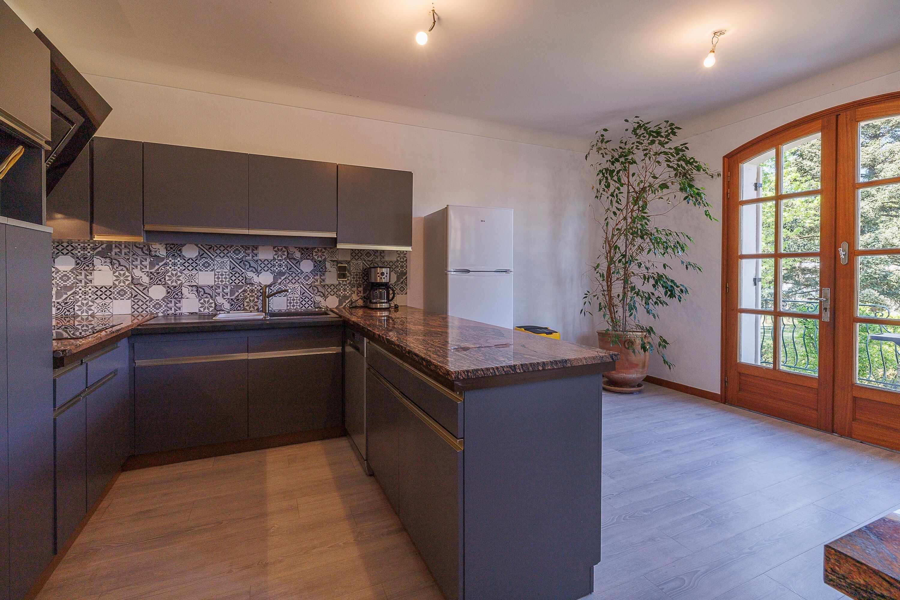 A modern kitchen with dark cabinets, a marble countertop, and a built-in oven. There is a potted plant and a large window with a view of greenery.