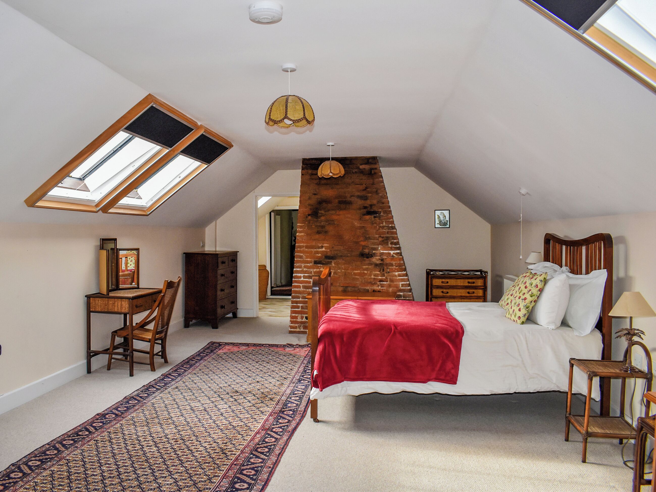 A bedroom with a sloped ceiling, a skylight, a bed with a red bedspread, a wooden dresser, a chair, and a patterned rug.
