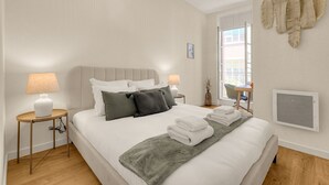 A neatly made bed with pillows and a blanket, a bedside table with a lamp, and a window with a view of buildings. - Bloom - 12min à pied du Capitole (Toulouse)