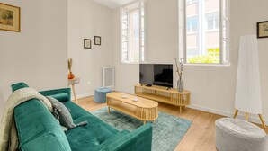 A modern living room with a green sofa, a wooden coffee table, and a TV on a wooden stand. There are two windows with white frames, a white easel, and a vase with dried plants on a side table. - Bloom - 12min à pied du Capitole (Toulouse)
