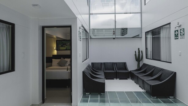Un moderno vestíbulo de hotel con un separador de vidrio, una zona de estar con sofás negros, una planta de cactus y una cama en la habitación contigua.