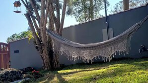 Un hamac est suspendu entre deux arbres dans un jardin, avec une maison en arrière-plan.