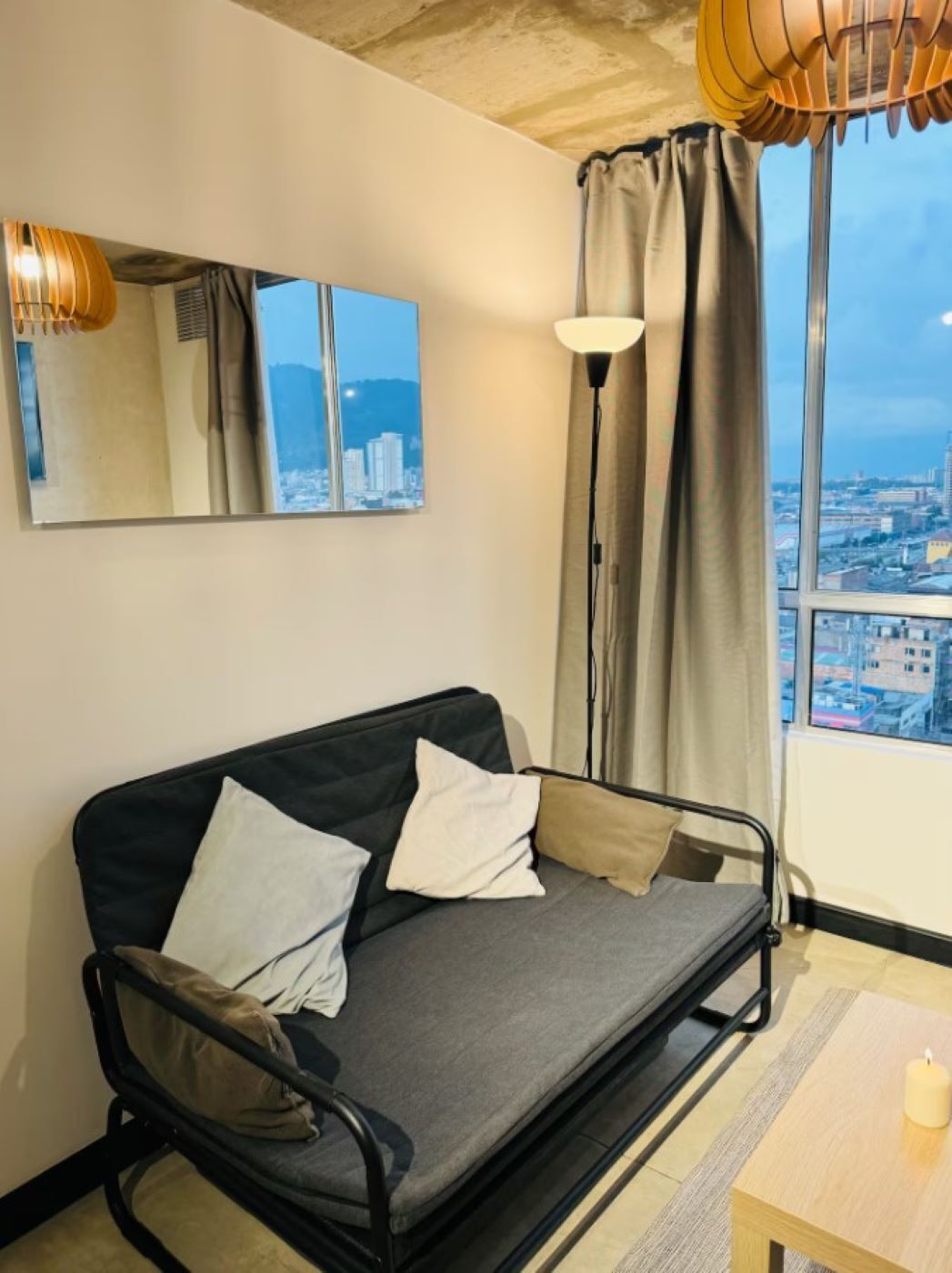 A compact room with a sofa bed, a small table, a floor lamp, and a cityscape view through the window.