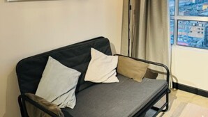 Une pièce compacte comprenant un canapé-lit, une petite table, une lampe sur pied et une vue sur le paysage urbain par la fenêtre.