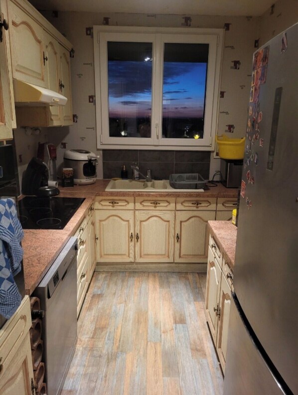 A kitchen with wooden cabinets, a stainless steel refrigerator, a window with a view of the sunset, and a sink. - Private room (Allonnes)