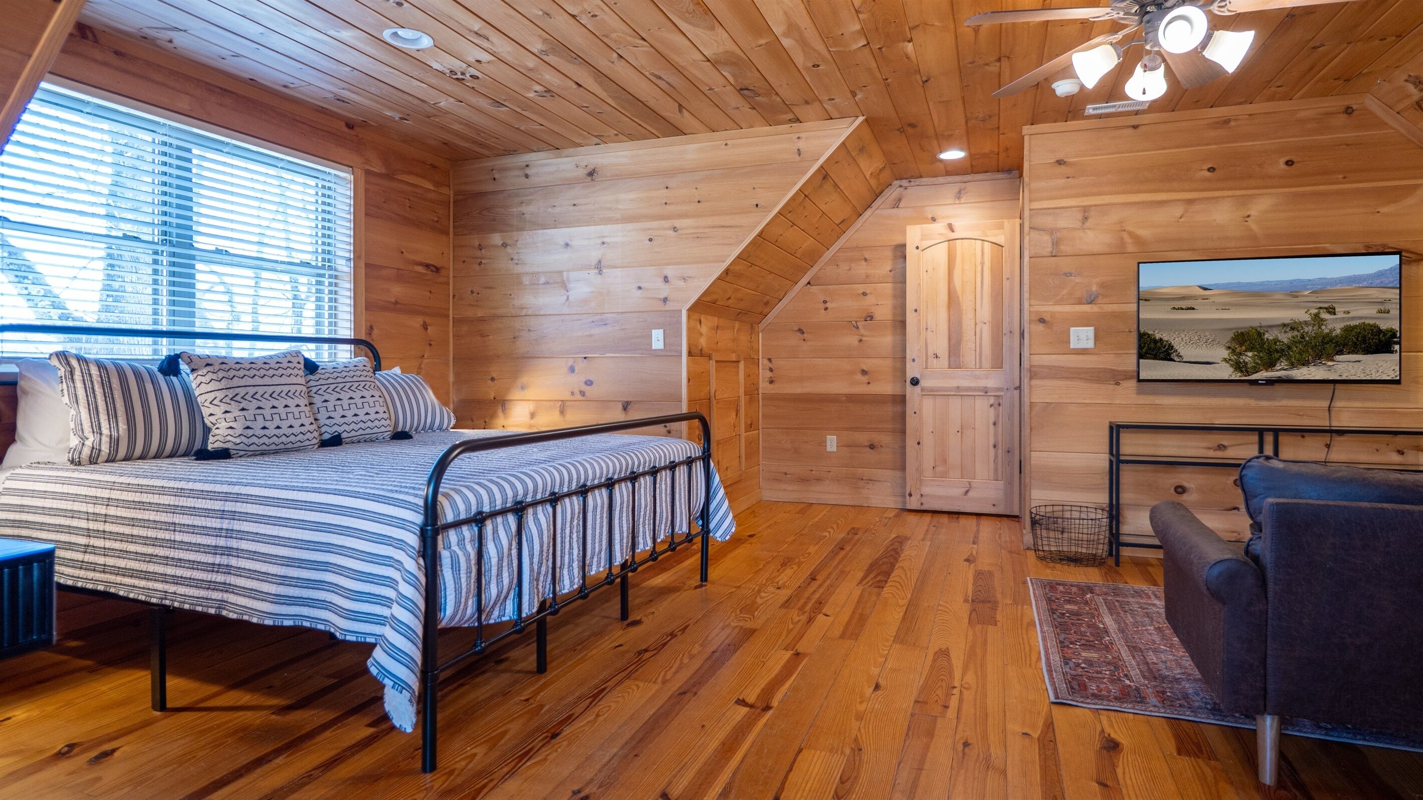 A cozy wooden cabin room with a bed, a chair, a television, and a window with blinds.