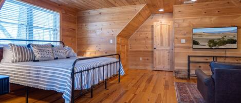A cozy wooden cabin room with a bed, a chair, a television, and a window with blinds.