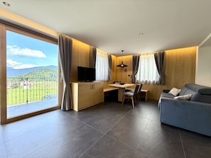 Een moderne woonkamer met een grote schuifpui naar een balkon, een houten bureau met een televisie, een bank en een eettafel met stoelen.