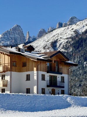 A two-story wooden chalet with balconies, set against a backdrop of snow-covered mountains.