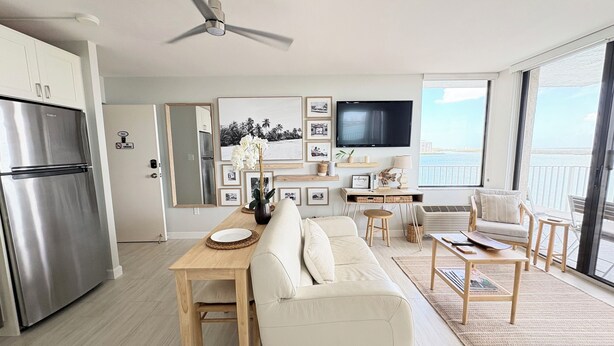 A modern kitchen with a built-in refrigerator, a dining area with a wooden table and white sofa, and a living area with a TV and a balcony with a view of the sea.
