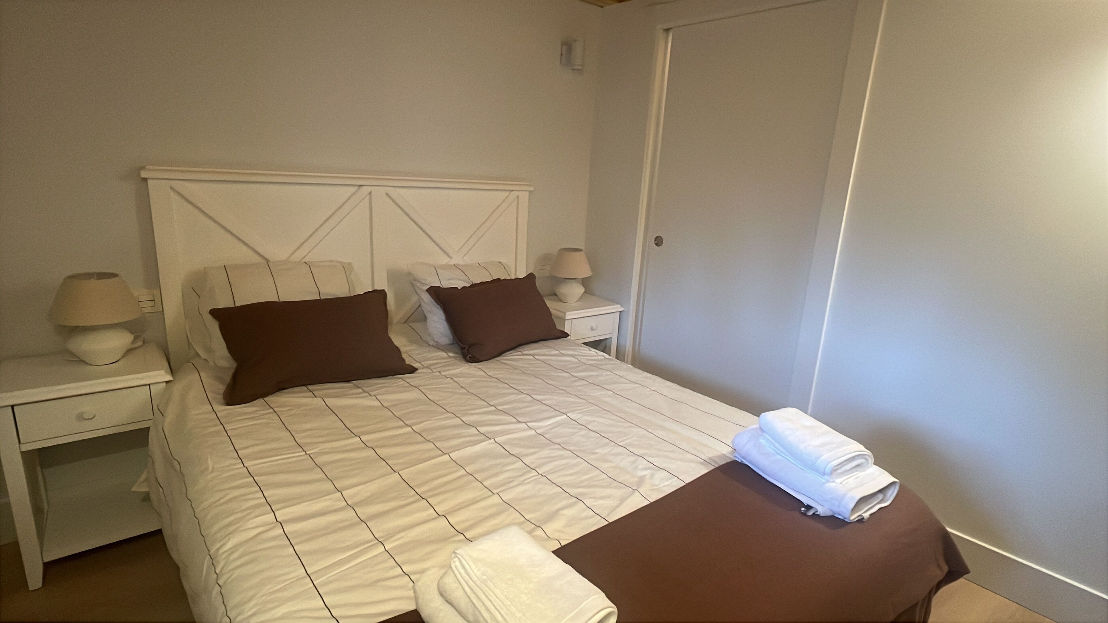 A neatly made bed with a white and brown checkered bedspread, two pillows, and a folded towel on the side. There are two bedside tables with lamps and a nightstand with a lamp.