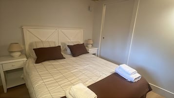 A neatly made bed with a white and brown checkered bedspread, two pillows, and a folded towel on the side. There are two bedside tables with lamps and a nightstand with a lamp.