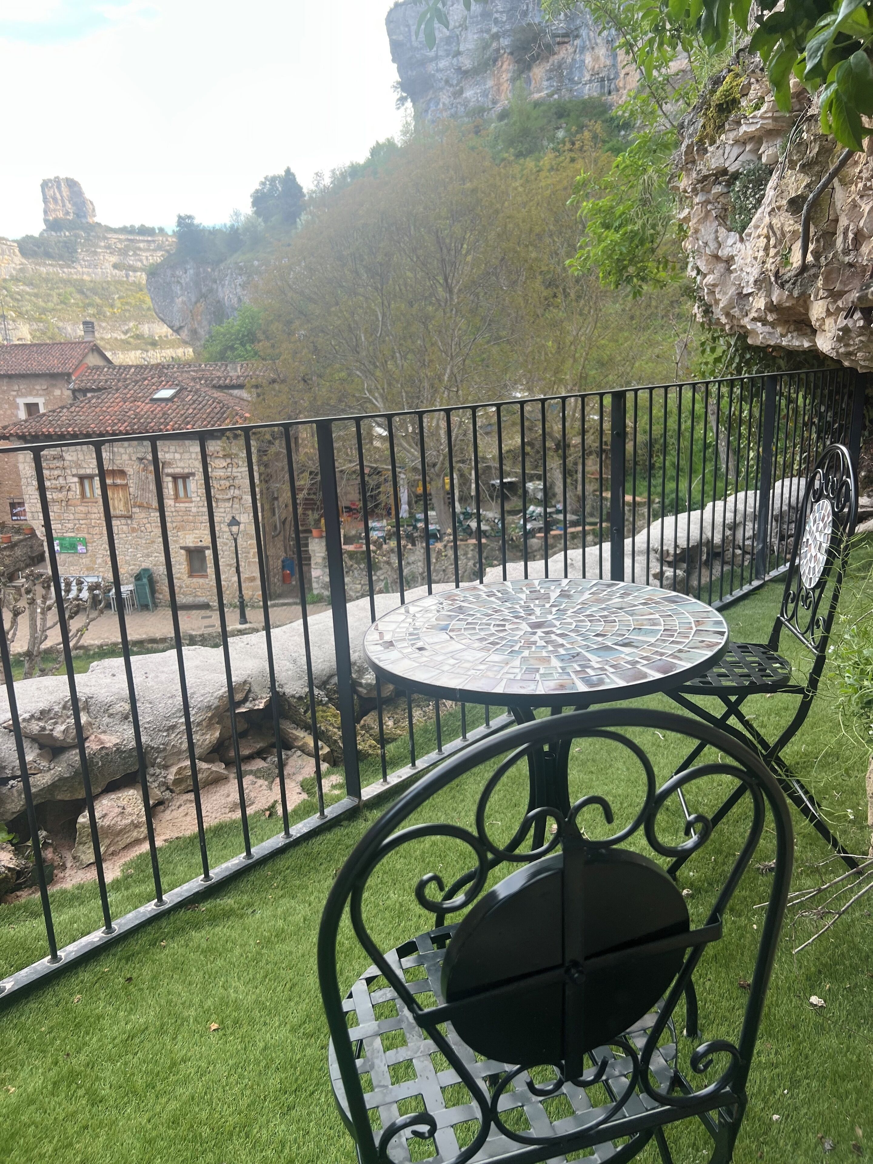 Ein Balkon mit einem Metalltisch und Stuhl, Blick auf eine felsige Klippe und ein Gebäude mit Ziegeldach im Hintergrund.