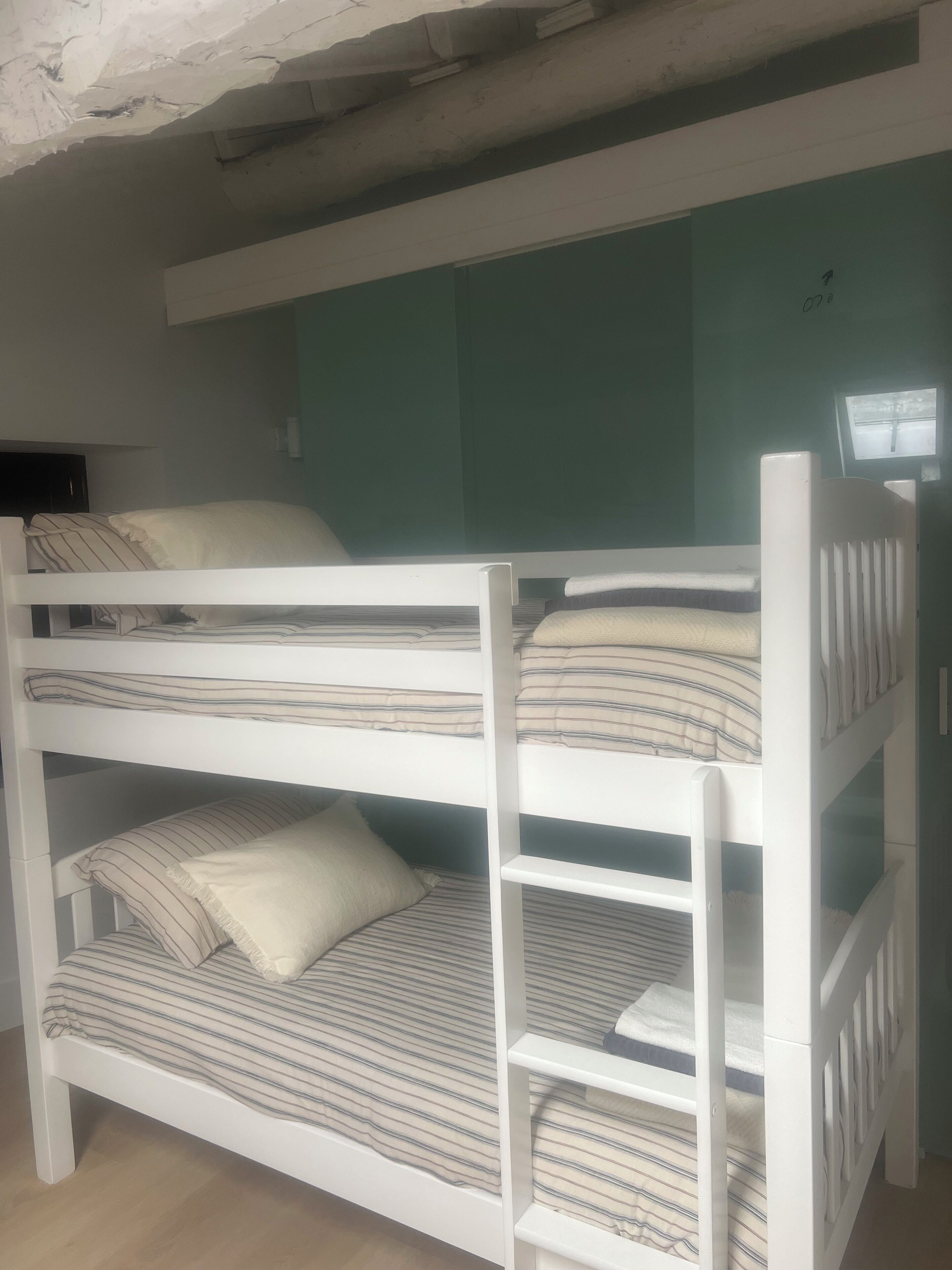 Una cama litera con ropa de cama a rayas y almohadas, un cabecero blanco y una ventana pequeña con vistas a una carretera.