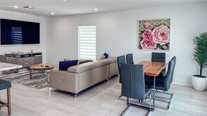 A modern living room with a sofa, dining table, and chairs, a flat-screen TV, and a large floral painting. - 664 Naples Beach Paradise-Your Next Vacation Spot with Private Pool & Spa (Naples)