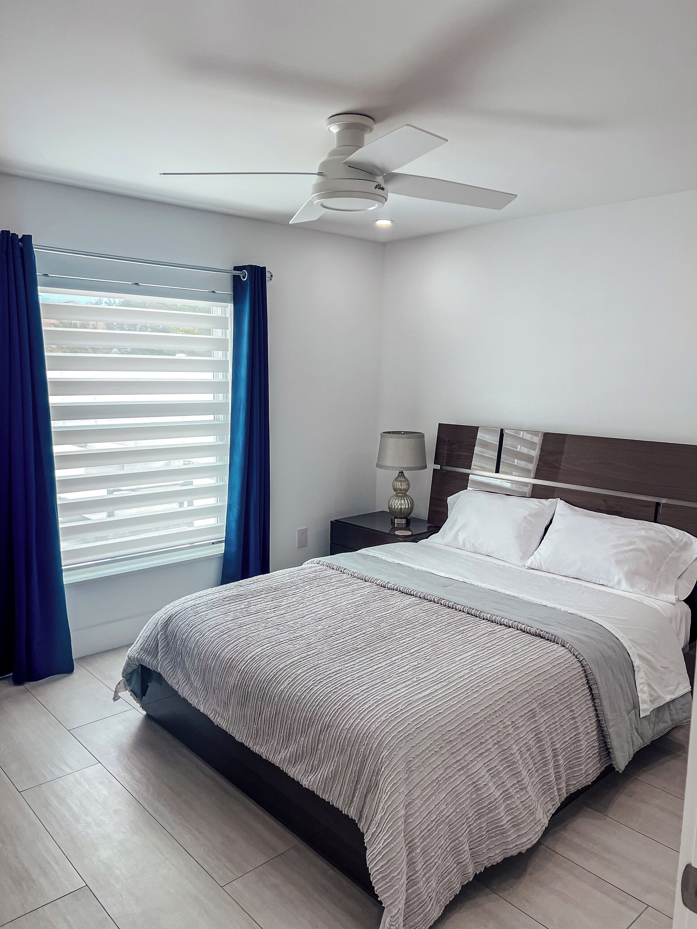 A bedroom with a bed, a nightstand, a lamp, a ceiling fan, and a window with blinds.