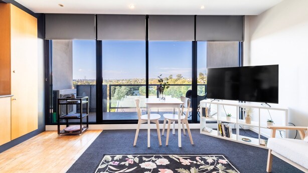 A modern living room with a large window, a flat-screen TV, a dining area with a table and chairs, and a bookshelf.
