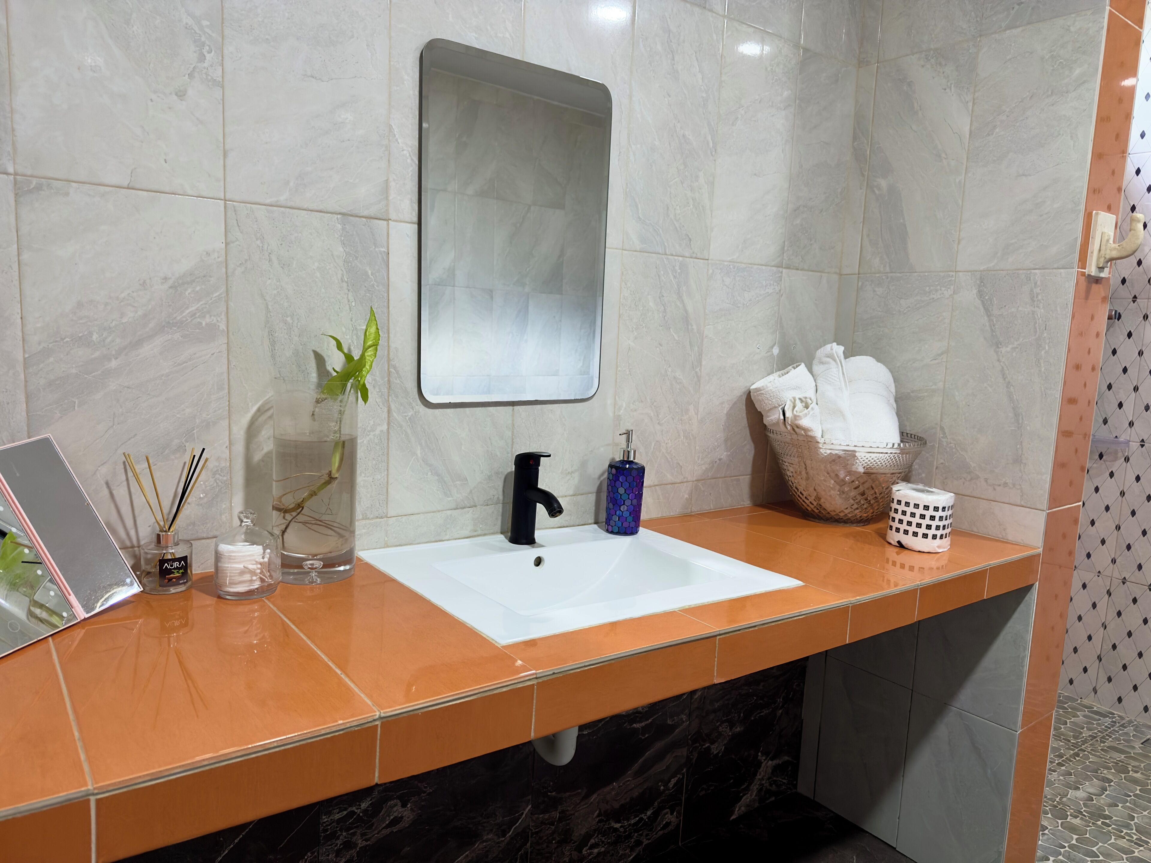 A bathroom with a white sink, black faucet, and a mirror above it. There is a plant in a glass container on the counter, a bottle of soap, and a basket with towels.
