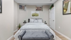 Un dormitorio con cama, mesitas de noche, un banco, una ventana y una puerta.