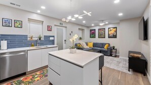 Una cocina moderna con armarios blancos, lavavajillas de acero inoxidable y una isla central. En la zona de estar, hay un sofá gris con cojines amarillos y un televisor de pantalla plana montado en la pared.
