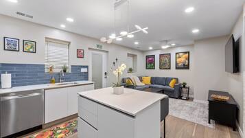 A modern kitchen with white cabinets, a stainless steel dishwasher, and a central island. There is a gray sofa with yellow cushions in the living area, and a flat-screen TV mounted on the wall.