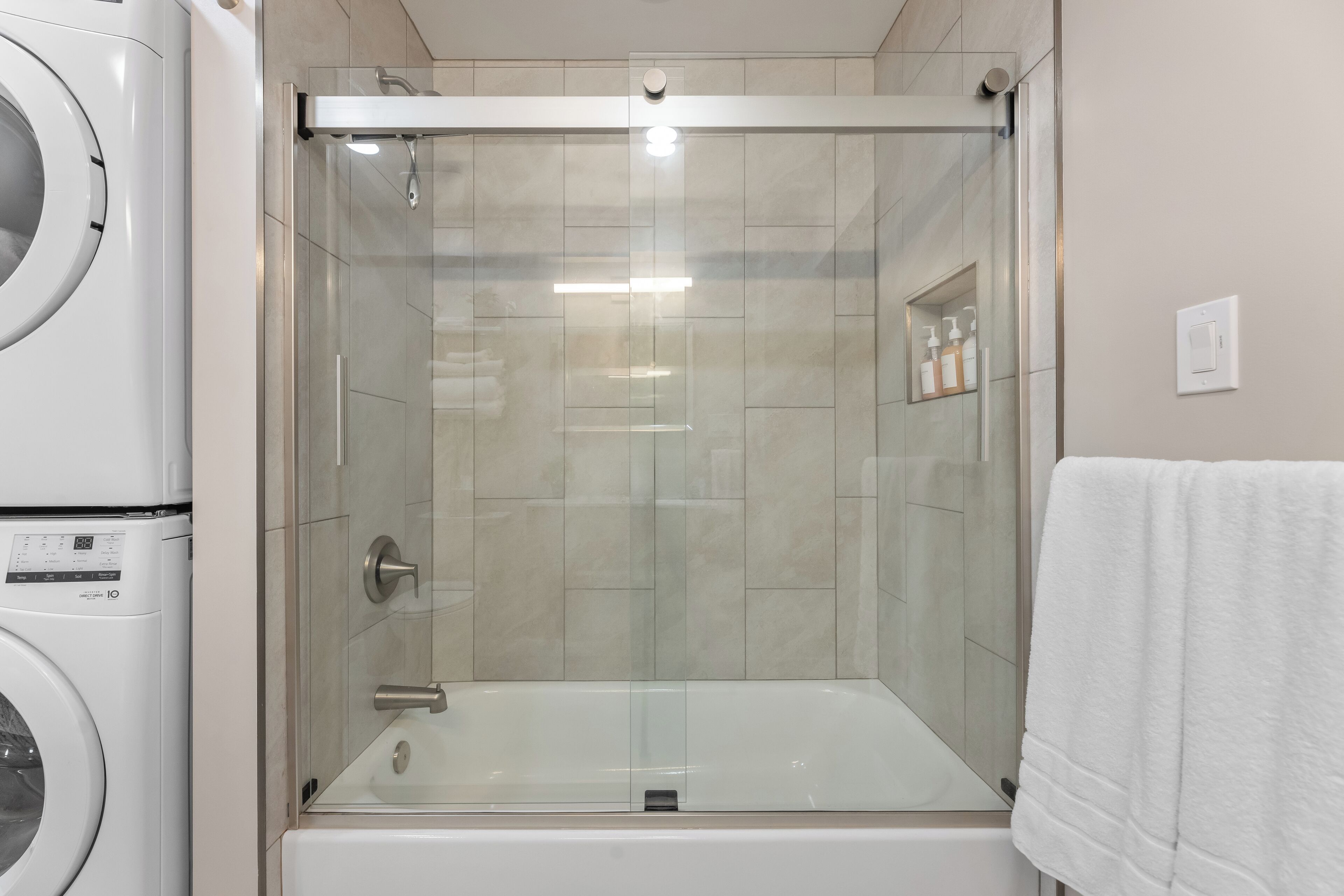 Ein modernes Badezimmer mit einer gläsernen Duschkabine, einer weißen Badewanne und einer gestapelten Waschmaschine und einem Trockner.