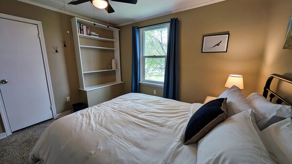 Un dormitorio con una cama, una mesita de noche, una ventana con cortinas, un ventilador de techo y una estantería.
